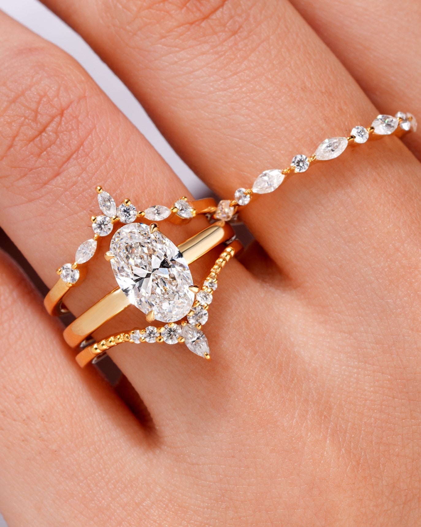 A close-up of a hand wearing a yellow gold ring set featuring a large oval-cut diamond center stone with smaller marquise and round diamonds, accompanied by a matching chain bracelet.